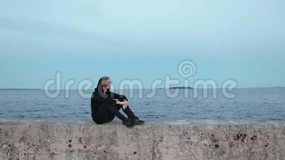 一个穿着黑色衣服的年轻人的肖像他坐在靠海的码头上放松视频的预览图