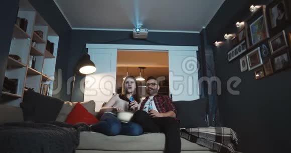 快乐的年轻放松的白种人浪漫的夫妇笑着在家看有趣的电影用投影仪慢动作吃零食视频的预览图