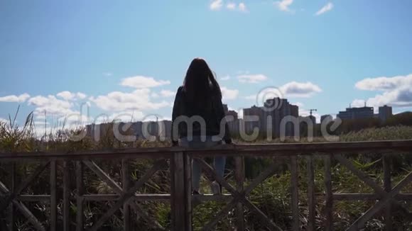 年轻女孩或穿绿色夹克的女人坐在栏杆上举起她的手城市湖泊和草地的背景慢慢视频的预览图