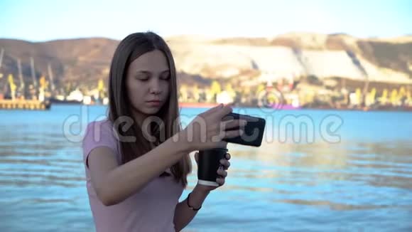 风姿绰约的年轻女子一边喝着咖啡一边用手机在山峦密布的河堤上自拍视频的预览图