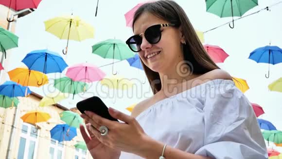 美丽的女游客在彩伞装饰的街道上使用手机视频的预览图
