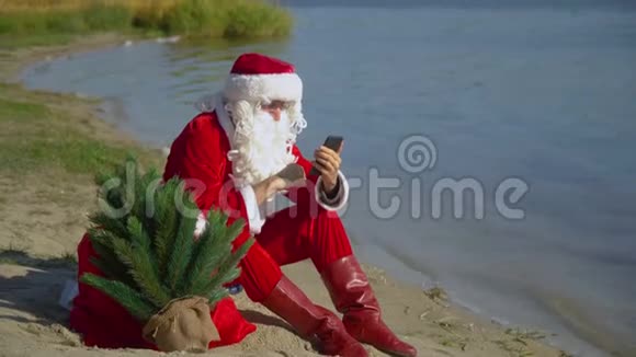 圣诞老人坐在湖边的沙滩上拿着一袋礼物在他的智能手机上工作视频的预览图