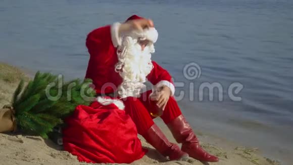圣诞老人坐在湖边的沙滩上嗅着酒精和瓶子的味道喝醉的圣诞老人视频的预览图