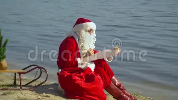 圣诞老人坐在湖边的沙滩上拿着一袋礼物在他的礼物清单上反思度假村的圣诞老人视频的预览图