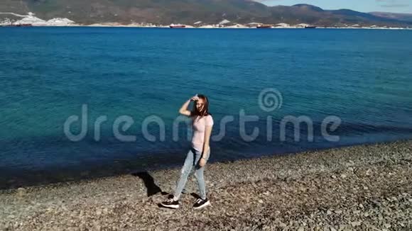穿着粉红色衬衫和蓝色破牛仔裤的女孩在海滩上散步在镜头中观看空中景观视频的预览图