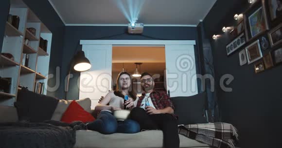 快乐的年轻白种人男人和女人坐在一起看有趣的电影在家里用零食使用投影仪慢动作视频的预览图
