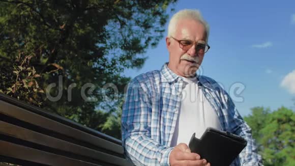 一个有胡子的老人正在读书中有趣的时刻老人坐在公园的长凳上看电子书视频的预览图