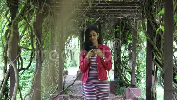 年轻的千禧一代女孩在花园里用电话穿过佩高拉或遮阳走道视频的预览图