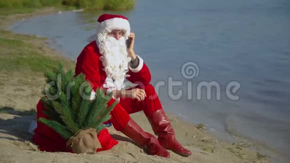 圣诞老人坐在湖边的沙滩上拿着一袋礼物用手机说话视频的预览图