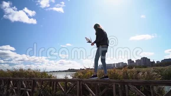 漂亮的女孩走在木栏杆上背景是湖芦苇和建筑物慢动作的快乐女人视频的预览图