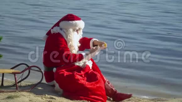 圣诞老人坐在湖边的沙滩上拿着一个礼品袋把一些东西写在清单上度假村的圣诞老人视频的预览图