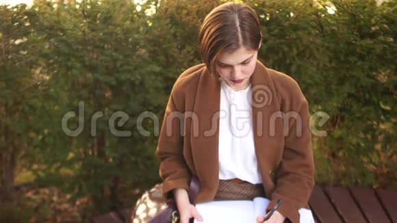 年轻的女学生在长凳上的公园里准备考试棕色外套短发笔记本和书籍视频的预览图
