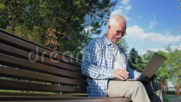 快乐老人在Skype上交谈坐在花园的长凳上一个养老金领取者把笔记本电脑放在膝上他是视频的预览图