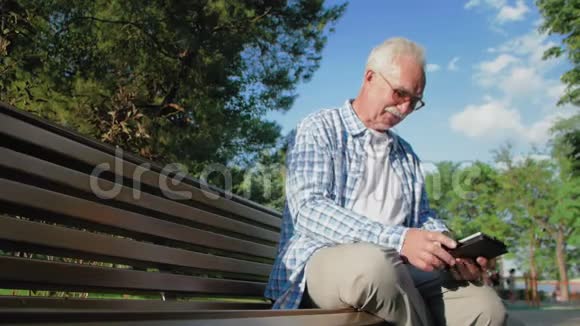 戴着眼镜和胡子的老人坐在公园的长凳上看电子书视频的预览图