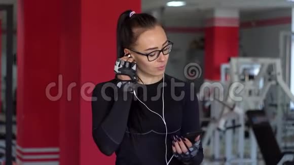 带着耳机的年轻女孩在健身房里用智能手机女孩听音乐视频的预览图