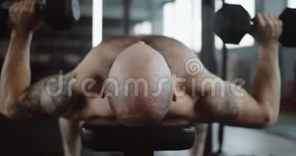 近距离后景年轻的高加索肌肉男躺在长凳上举哑铃在大型体育馆大厅慢动作视频的预览图