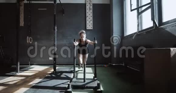 年轻漂亮的白人运动女性推着训练雪橇走向相机在硬体健身房锻炼慢动作视频的预览图