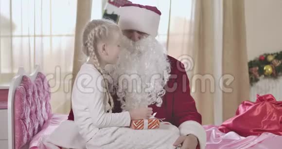 可爱的白种人女孩扎着辫子坐在圣诞老人的膝盖上在他的低语儿童在场视频的预览图