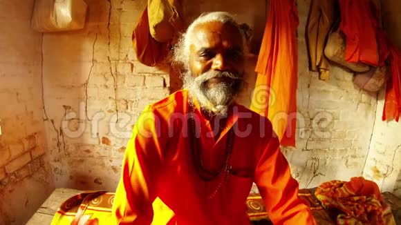 画像身穿橘色衣服脖子上挂着祈祷珠墙上挂着瓦工和衣服的老萨杜印度僧人视频的预览图