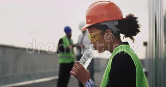 在建筑工地上一位口渴的非洲裔女士用塑料瓶饮水她戴着安全设备视频的预览图