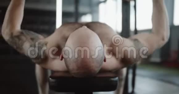 近距离后景年轻秃顶运动员举起沉重的哑铃在长凳上变得疲倦和休息在大型健身房视频的预览图