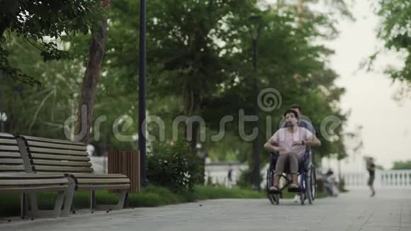 坐轮椅的快乐残疾人和在城市公园跑步的快乐年轻人视频的预览图