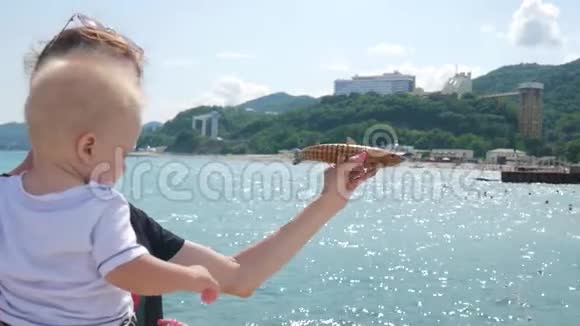 年轻的妈妈带着小儿子在大海的背景上玩玩具鱼视频的预览图