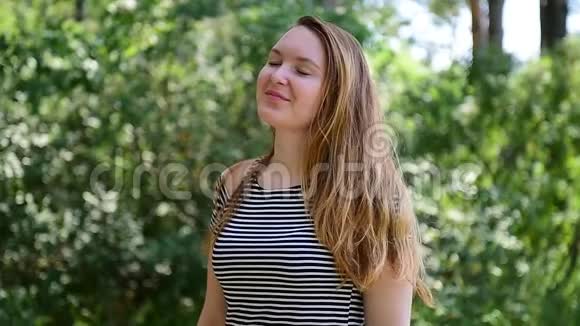 年轻的棕色头发的女人穿着条纹t恤站在阳光下迎着绿色的树叶风吹起她的头发漂亮姑娘视频的预览图