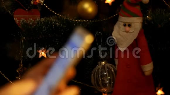 雌性手拿着一部智能手机在圣诞树的背景上关闭圣诞老人和手机视频的预览图