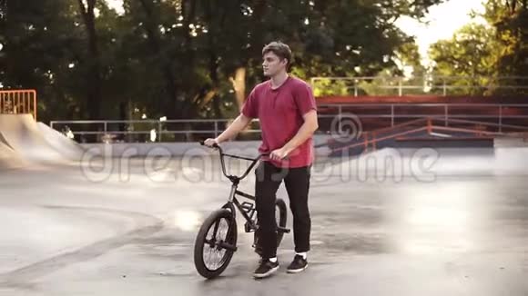 穿着红色衬衫和黑色裤子的白人男子走在现代滑板公园旁边有他的BMX自行车天气晴朗视频的预览图