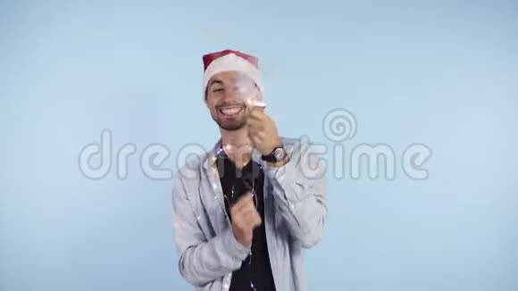 年轻的微笑的男人穿着休闲服头上戴着圣诞老人帽在蓝色背景上爆炸了纸屑饼干快乐视频的预览图