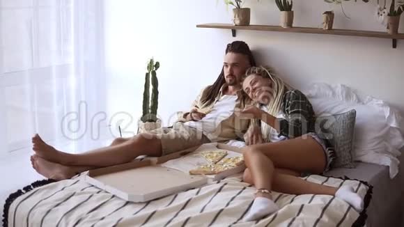 可爱的一对可怕的夫妇有一个懒惰的早晨吃送外卖披萨躺在床上看电视视频的预览图