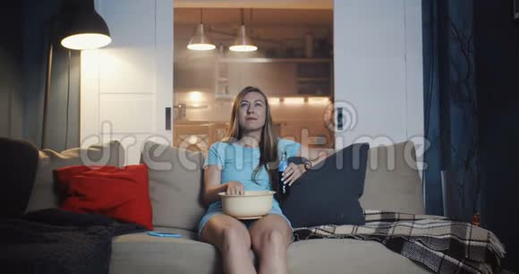 快乐的年轻美丽的随意的高加索女人坐在家里的沙发上用爆米花慢动作看电影视频的预览图