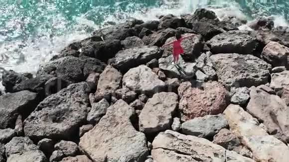 穿着红色漂亮衣服的女人双手分开站在岩石峭壁海滩上海浪拍打着岩石溅起水花视频的预览图