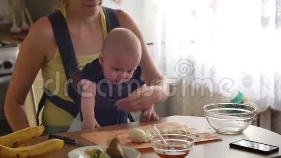 年轻女子带着一个蹒跚学步的男孩坐在吊带里坐在厨房的桌子旁切鸡蛋完成后视频的预览图