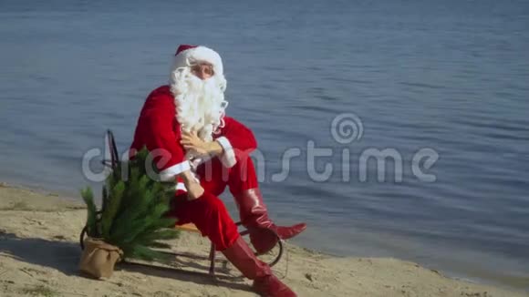 圣诞老人坐在沙滩上的雪橇上把他的胡子眼镜和帽子固定在镜头里视频的预览图