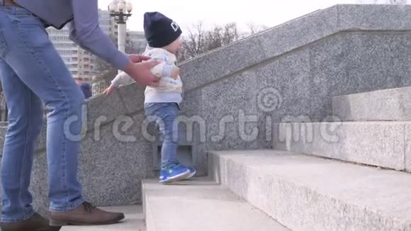 戴墨镜的大胡子牵着他的小儿子的手帮助小男孩爬上城市公园的楼梯视频的预览图