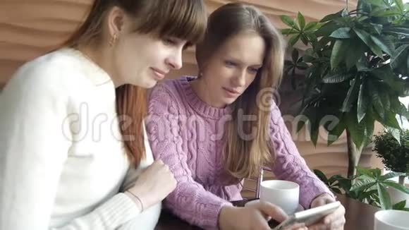 两个年轻的女性朋友正在智能手机上的社交媒体上看照片开心地微笑着在咖啡上交谈视频的预览图