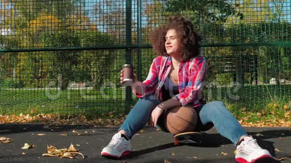 一个十几岁的小女孩坐在操场上的滑板上喝着纸杯里的咖啡拿着篮球视频的预览图