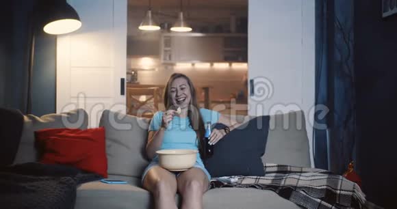 快乐年轻迷人的欧洲女人微笑享受在家里看电影的零食和喝慢动作视频的预览图