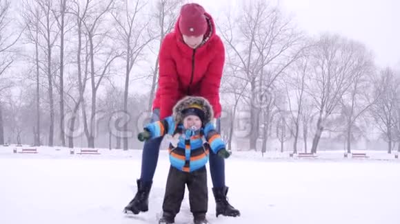 穿着红色羽绒服的年轻女子在冬天的城市公园背景下在雪地里缓慢地把她的小儿子卷在冰上视频的预览图