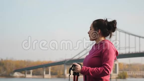 可爱的胖乎乎的女人在密集的运动前从城市河边和公园的瓶子里喝水拿着杆子在北欧散步视频的预览图