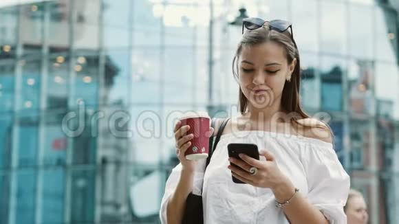 微笑可爱的女人带着一杯咖啡在商业街使用手机视频的预览图