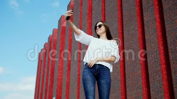 美丽的女人用手机在一座现代化的砖楼附近自拍视频的预览图