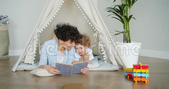 漂亮的女孩在公寓里给躺在帐篷里的聪明的孩子读书视频的预览图