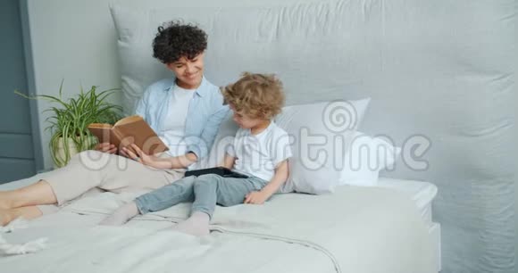 年轻的女人和小男孩在床上用平板电脑微笑着一起享受小玩意视频的预览图