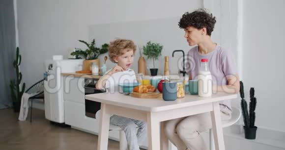 一位年轻的女士和一个小男孩在家厨房里吃麦片和牛奶微笑着视频的预览图