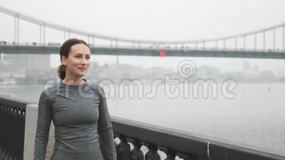 喜笑颜开的女人穿着活动服装在城市的堤岸上伸展背景是混凝土桥年轻女运动员担架视频的预览图