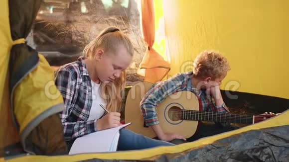 十几岁的男孩和女孩带着吉他坐在森林里的露营帐篷里少女记笔记的思考视频的预览图