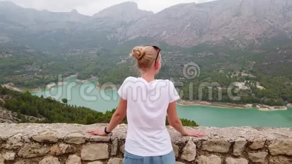 一个年轻的女孩从一座古老的城堡的墙上看到一个美丽的风景有一个蓝色的湖和山视频的预览图